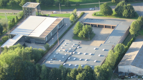 Luftaufnahme des DRK Katastrophenschutzzentrums Mönchengladbach Güdderath: Ein flacher, rechteckiger Industrie-/Bürokomplex mit mehreren Flachdächern und Dachaufbauten, umgeben von dichtem Baumbestand und Rasenflächen; im Hintergrund verläuft oben im Bild eine Straße mit geparkten Autos.